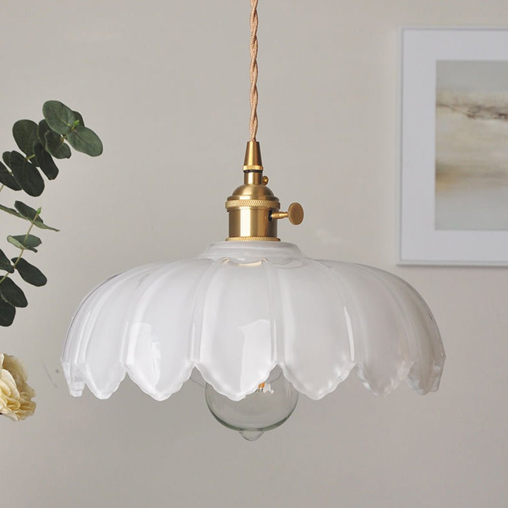 Een elegante hanglamp met een transparante glazen kap die een speelse, golvende vorm heeft. De lamp is bevestigd aan een gouden fitting met een gedraaide kabel, waardoor een chique vintage uitstraling ontstaat. De zichtbare lichtbron zorgt voor een warme gloed en creëert een uitnodigende sfeer in de ruimte, terwijl de groene plant op de achtergrond de stijl van de lamp accentueert.